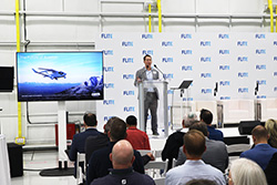 Man speaking at a podium in front of a seated audience with a screen showing an aerial photo
