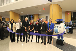 Group photo of airport staff and the mascot in front of a ribbon at a gate area
