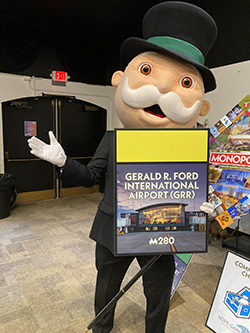 Monopoly Man-costumed mascot posing with a Gerald R. Ford International Airport board space sign