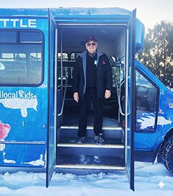 Man posing in the open doorway of a blue airport shuttle bus in the snow